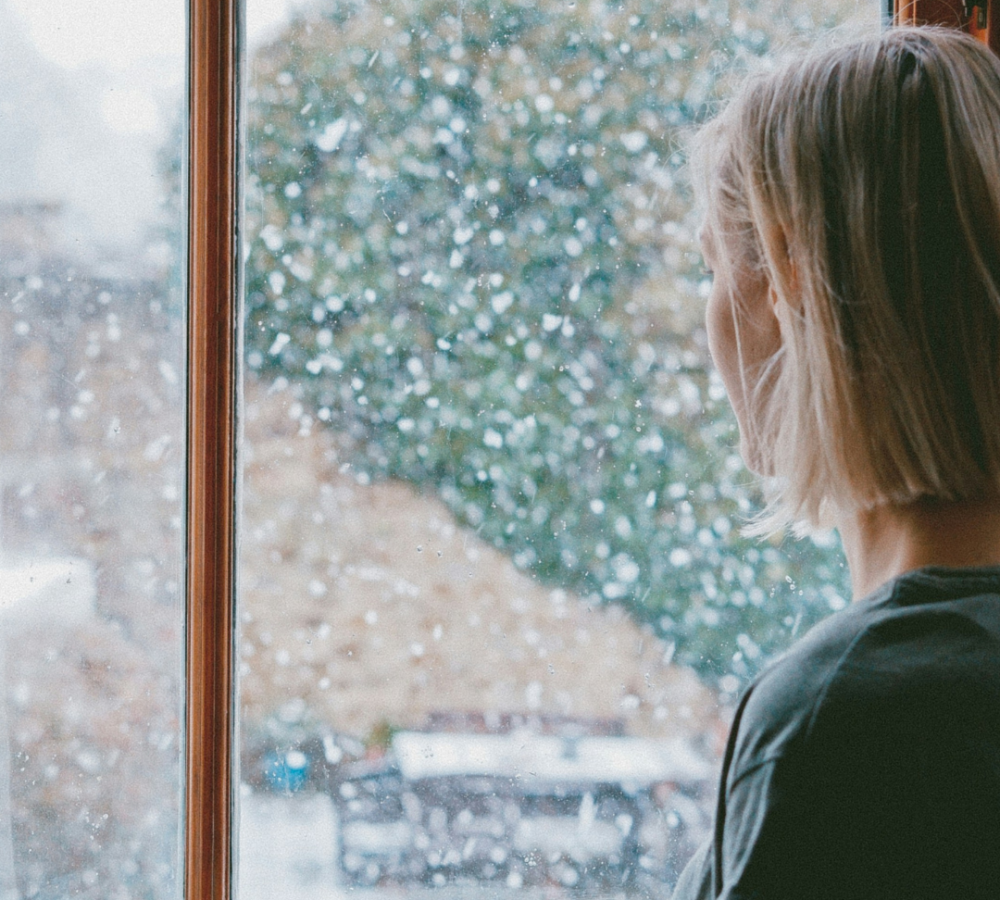 Woman standing by a window watching snow fall, appearing thoughtful and sad, suggesting grief during the holiday season.