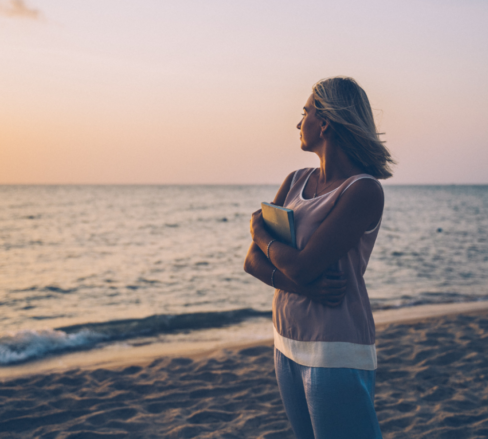 Woman standing on a beach at sunset, holding a journal and looking out at the ocean, symbolizing emotional healing and reflection after abortion.
