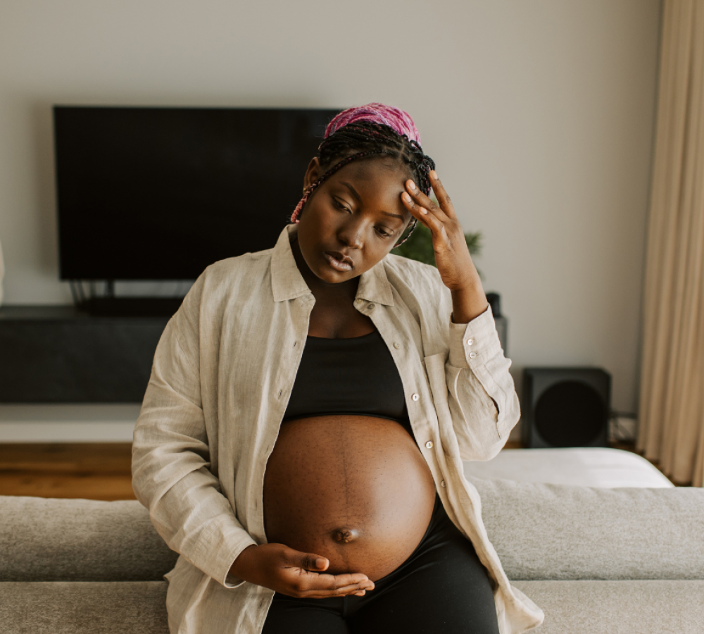 A visibly pregnant woman sits on a couch, resting one hand on her belly and the other on her forehead, appearing overwhelmed and deep in thought. Her expression suggests emotional strain, highlighting a need for support during pregnancy.