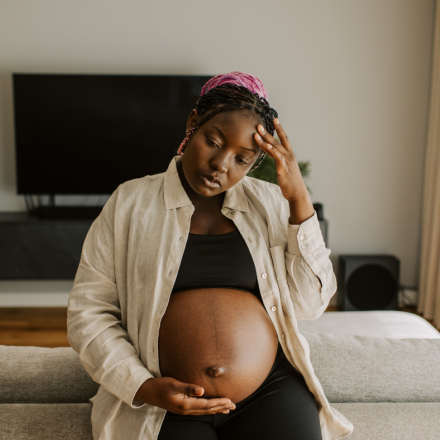 A visibly pregnant woman sits on a couch, resting one hand on her belly and the other on her forehead, appearing overwhelmed and deep in thought. Her expression suggests emotional strain, highlighting a need for support during pregnancy.