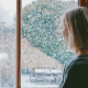 Woman standing by a window watching snow fall, appearing thoughtful and sad, suggesting grief during the holiday season.