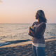 Woman standing on a beach at sunset, holding a journal and looking out at the ocean, symbolizing emotional healing and reflection after abortion.