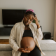 A visibly pregnant woman sits on a couch, resting one hand on her belly and the other on her forehead, appearing overwhelmed and deep in thought. Her expression suggests emotional strain, highlighting a need for support during pregnancy.